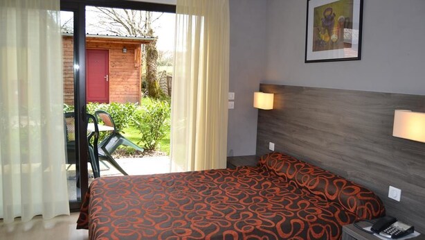 A hotel room with a bed, a nightstand with a phone, a wall-mounted lamp, and a view of a garden through a sliding door.