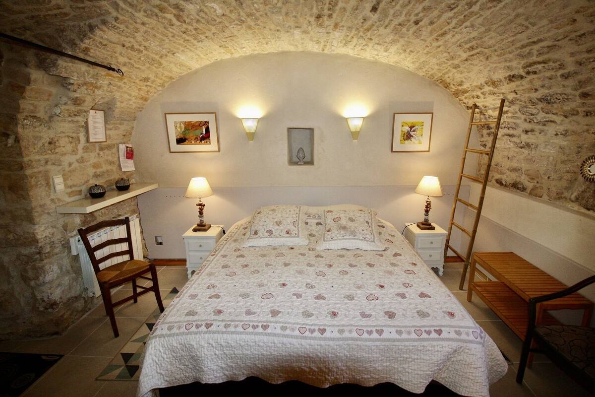 Un dormitorio con pared de piedra, cama con edredón floral, dos mesitas de noche con lámparas, una silla de madera y una pequeña mesa de madera.