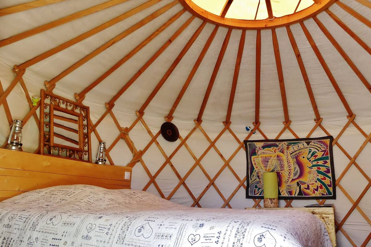 Interior de una yurta con cama, vigas de madera, una lámpara colgante y un cuadro colorido.