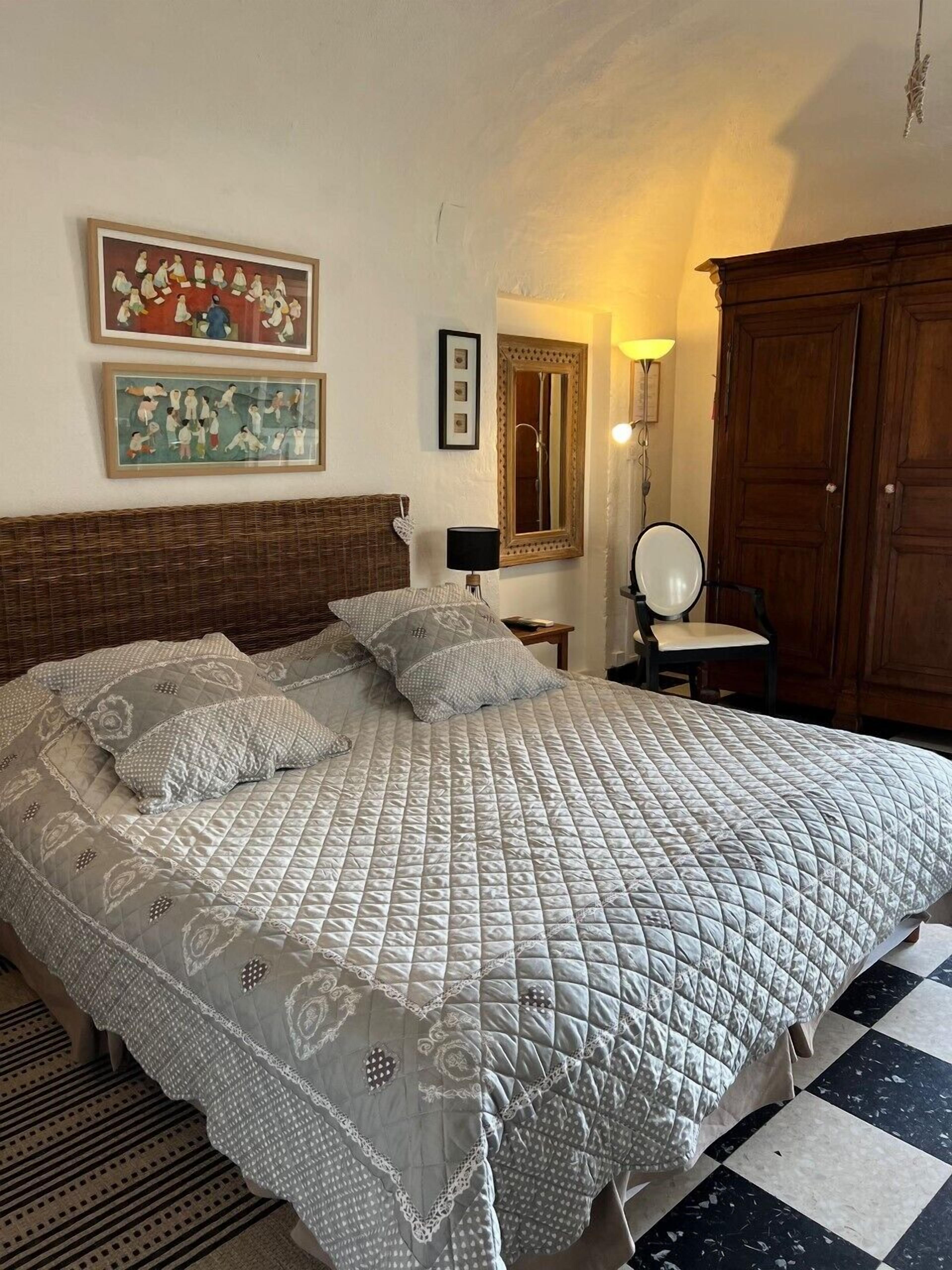 Una cama bien hecha con cubrecama acolchado, cabecera de madera y suelo a cuadros. En la pared hay cuadros enmarcados y un espejo sobre la cama.