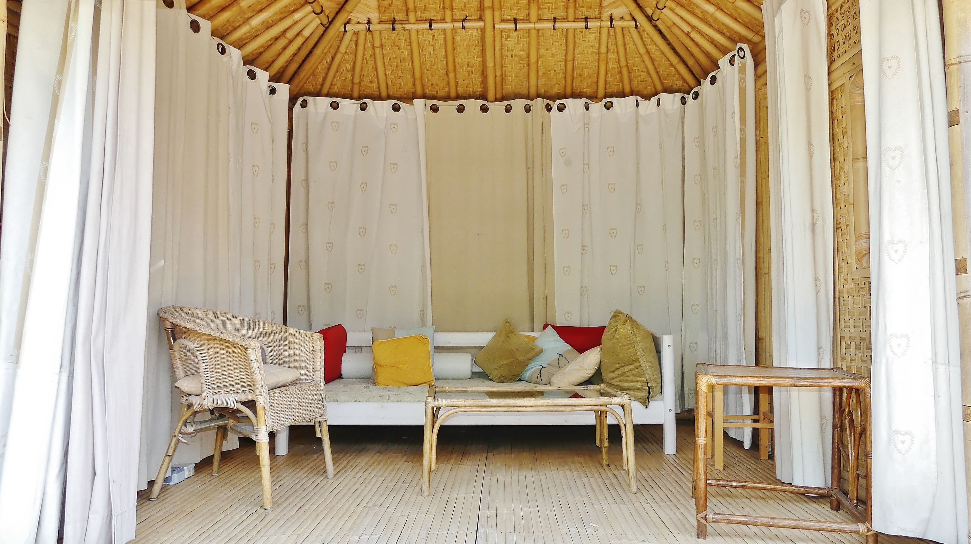 A room with a woven ceiling, white curtains with heart patterns, a wicker sofa with cushions, and a small wooden table.
