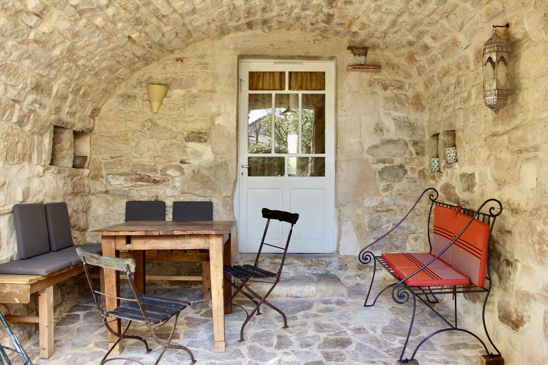 A stone-walled room with a wooden table and chairs, a bench, and a door with a window.