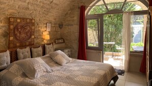 A bedroom with a stone wall, a bed with patterned bedding, a wooden headboard, a wall-mounted lamp, and a view of the outdoors through a glass door. - Mas Escombelle Chambre d'Hôte (Barjac)