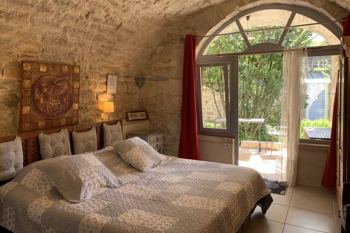 Un dormitorio con pared de piedra, cama con ropa de cama estampada, cabecero de madera, lámpara de pared y vistas al exterior a través de una puerta de cristal.