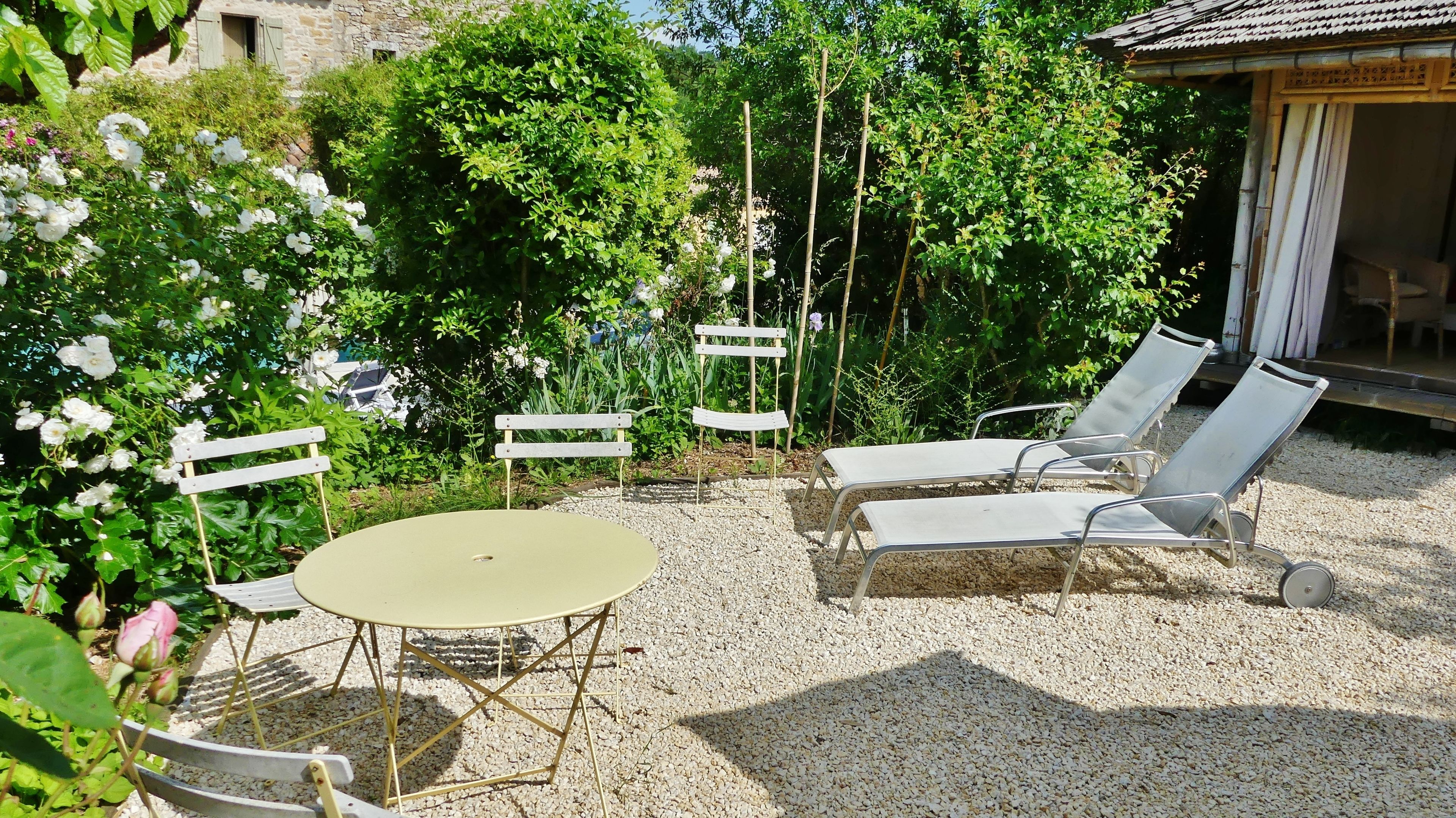 A patio with a round table and four lounge chairs, surrounded by gravel and lush greenery.
