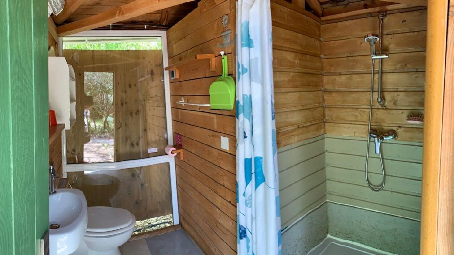 Un baño de madera pequeño y funcional con inodoro, ducha y una botella verde en la pared.