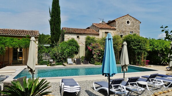 Un espace piscine agrémenté de chaises longues et de parasols, entouré de verdure et bordé d’un bâtiment en pierre doté d’un toit en tuiles.