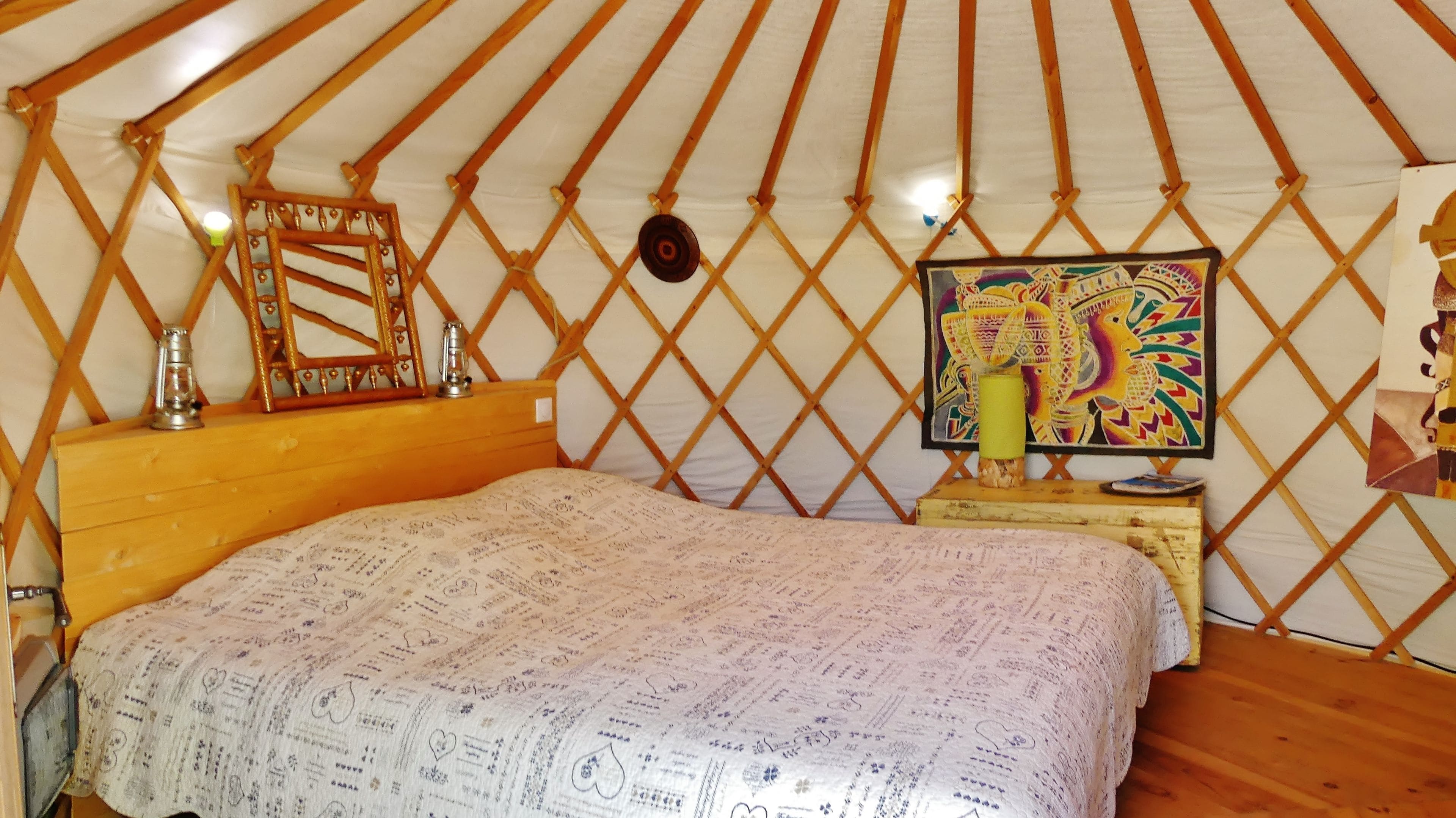 Una habitación acogedora con una cama, cabecera de madera, una mesita con una lámpara y un cuadro de colores en la pared.