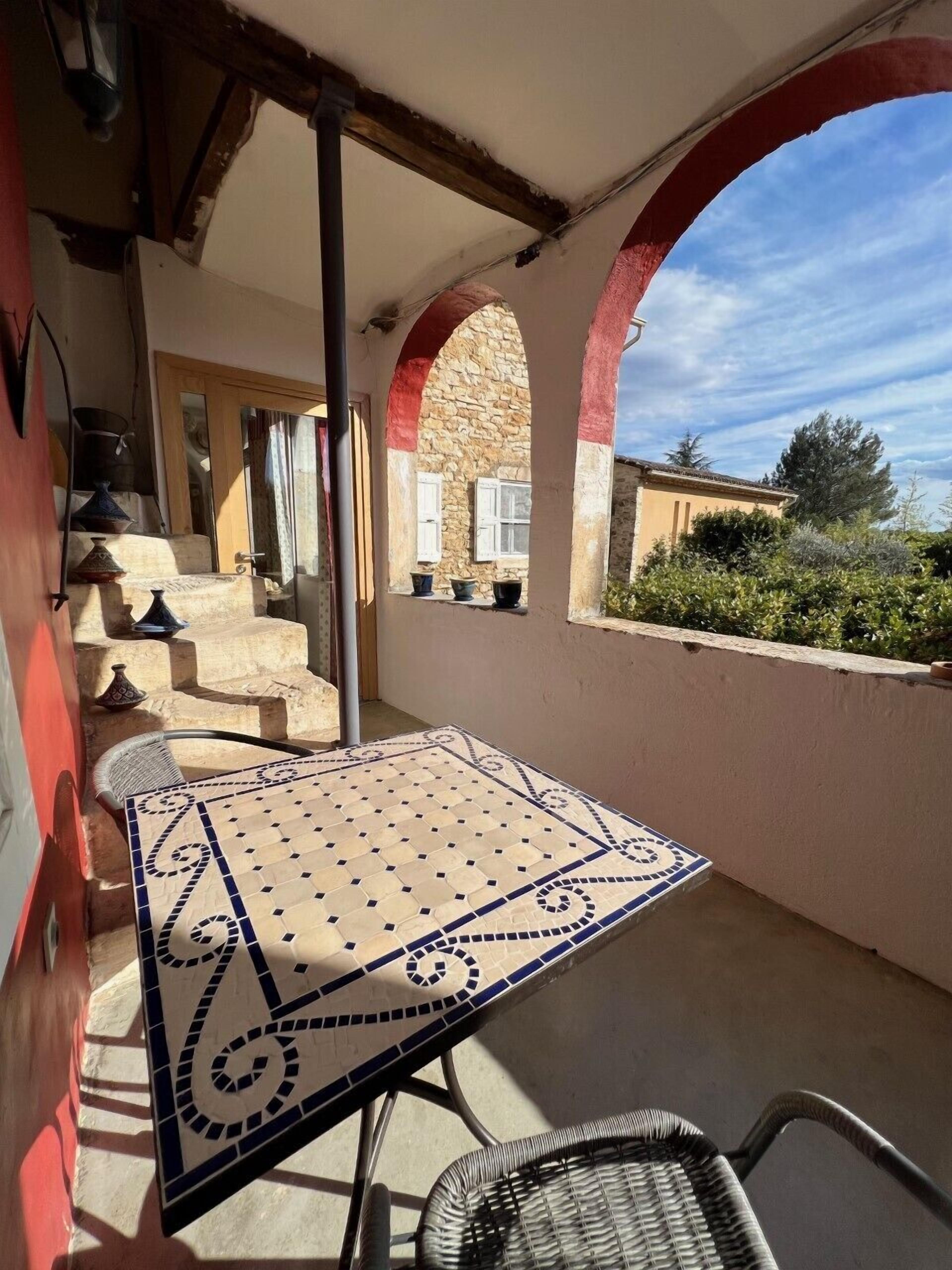 Uma varanda com mesa e cadeiras de azulejo, um muro de pedra e vista para um edifício com um arco vermelho.