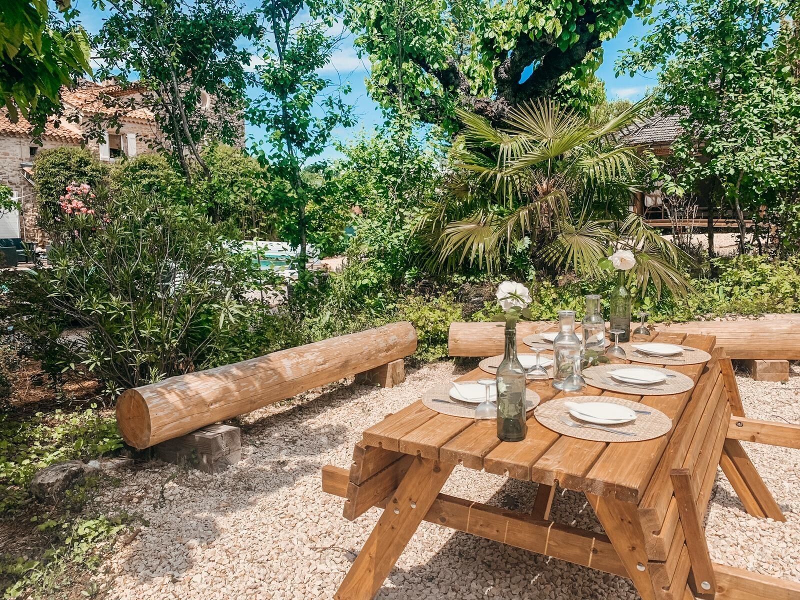 Ein Holztisch im Freien ist mit Tellern, Gläsern und einer Blumenvase gedeckt, umgeben von Grünpflanzen und einer Steinmauer.