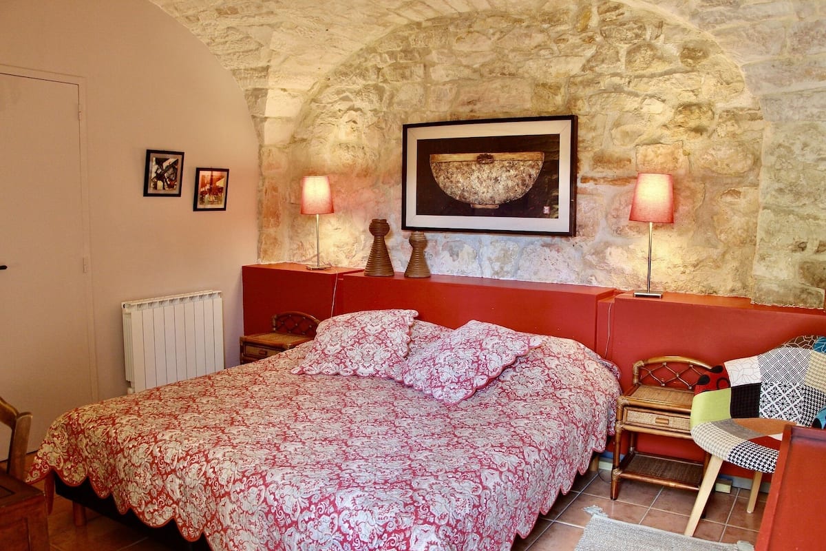 Un dormitorio con pared de piedra, una cama con colcha estampada, dos lámparas de noche, un cuadro enmarcado sobre la cama y una pequeña silla de madera.