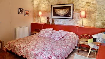 A bedroom with a stone wall, a bed with a patterned bedspread, two bedside lamps, a framed picture above the bed, and a small wooden chair.