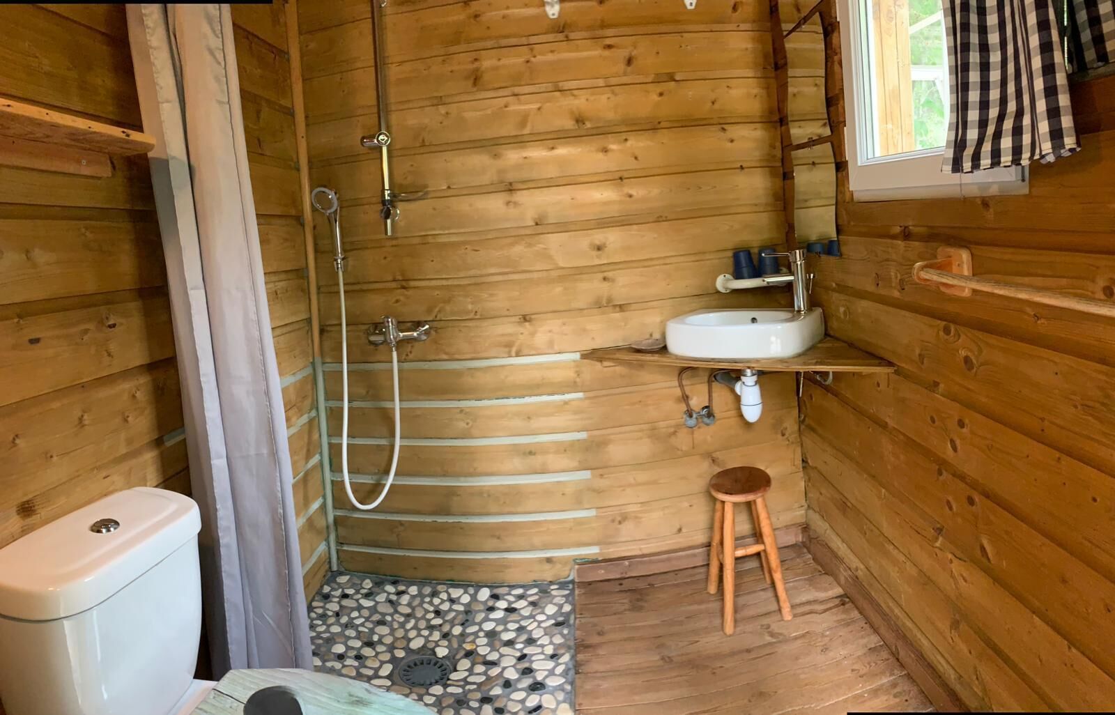 Bany d'una cabana de fusta amb dutxa, vàter, rentapla i un petit tamboret.