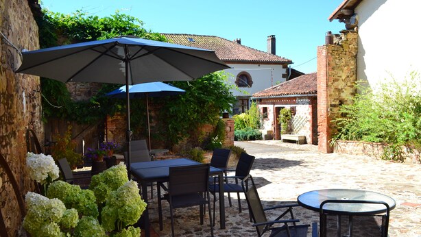 Een terras buiten met een tafel, stoelen, een grote parasol en potplanten.