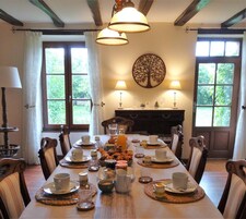 A dining room with a long table set for breakfast, surrounded by chairs, with a fireplace and a decorative tree wall hanging.