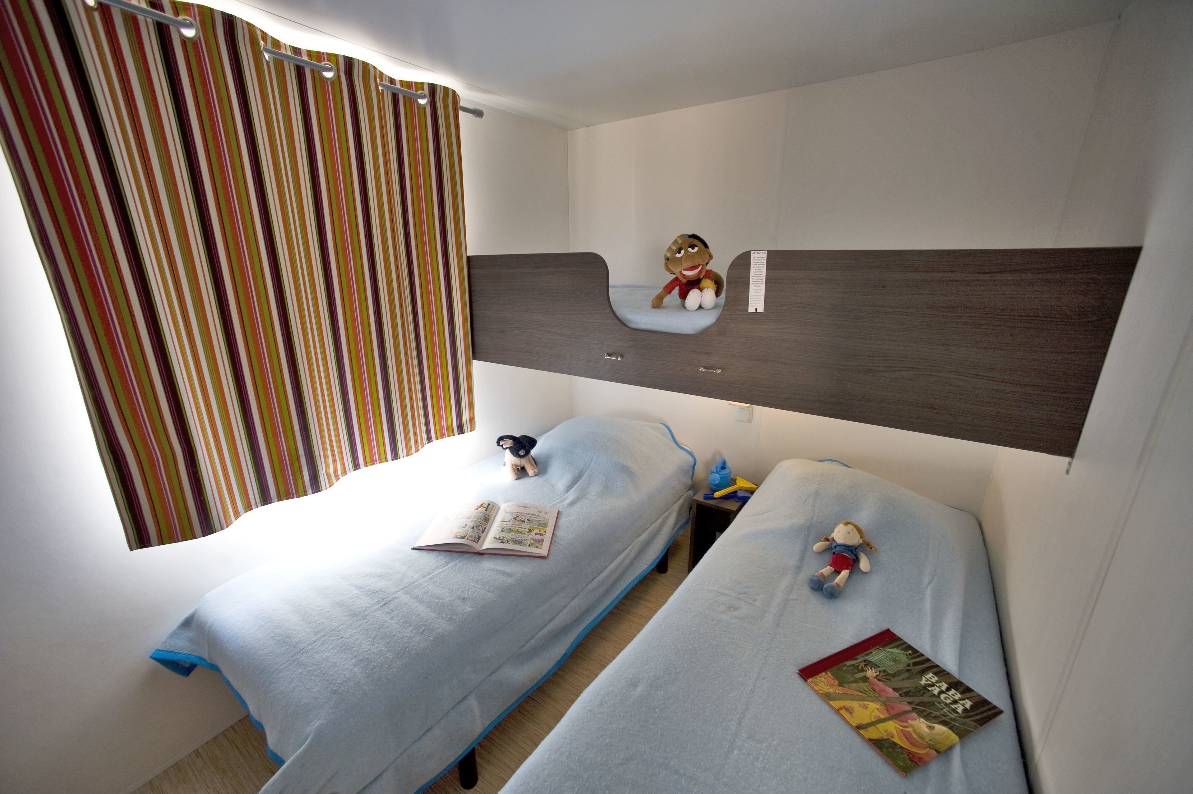 A room with two single beds, a striped curtain, a small shelf with a plush toy, and a book on one bed.