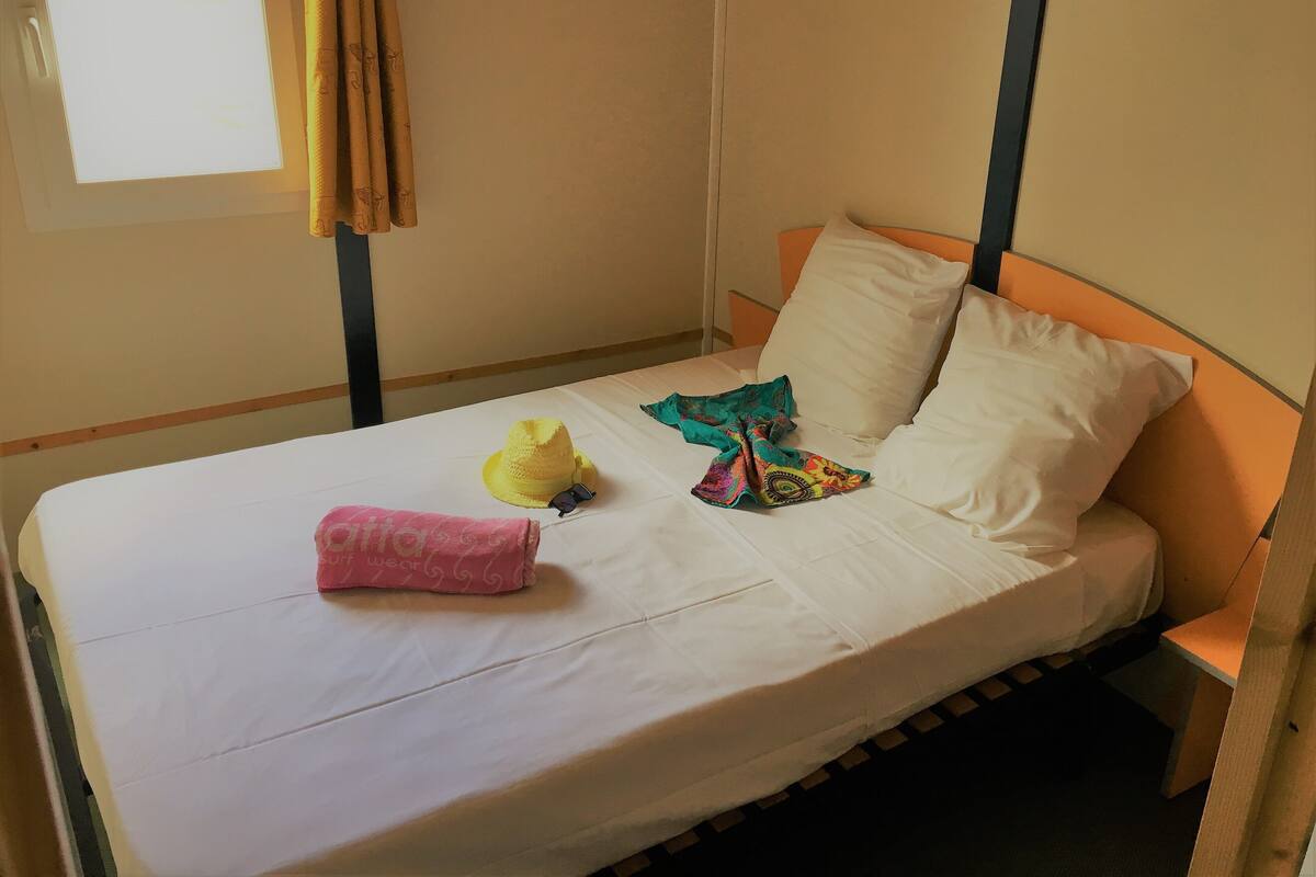 A single bed with white linens, a yellow hat, sunglasses, and a pink towel on the bed.