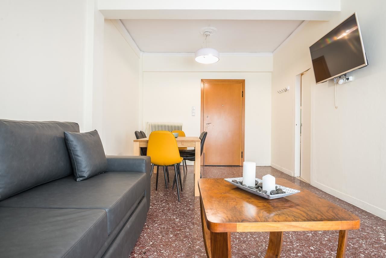 A compact room with a grey sofa, a wooden table with candles, a TV mounted on the wall, and a dining area with yellow chairs.