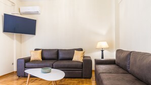 A living room with two sofas, a small white table, a lamp, and a mounted TV. - Victoria Queens Paradise Apartments (Athens)