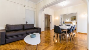 A living room with a dark brown sofa, a small white table, and a dining area with a white table and black chairs. - Victoria Queens Paradise Apartments (Athens)