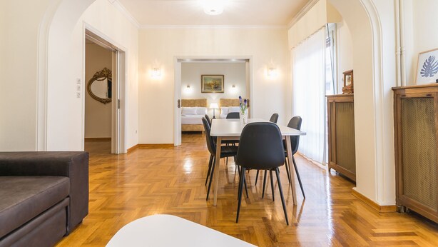 A dining area with a white table and black chairs, a sofa, and a fireplace. - Victoria Queens Paradise Apartments (Athens)