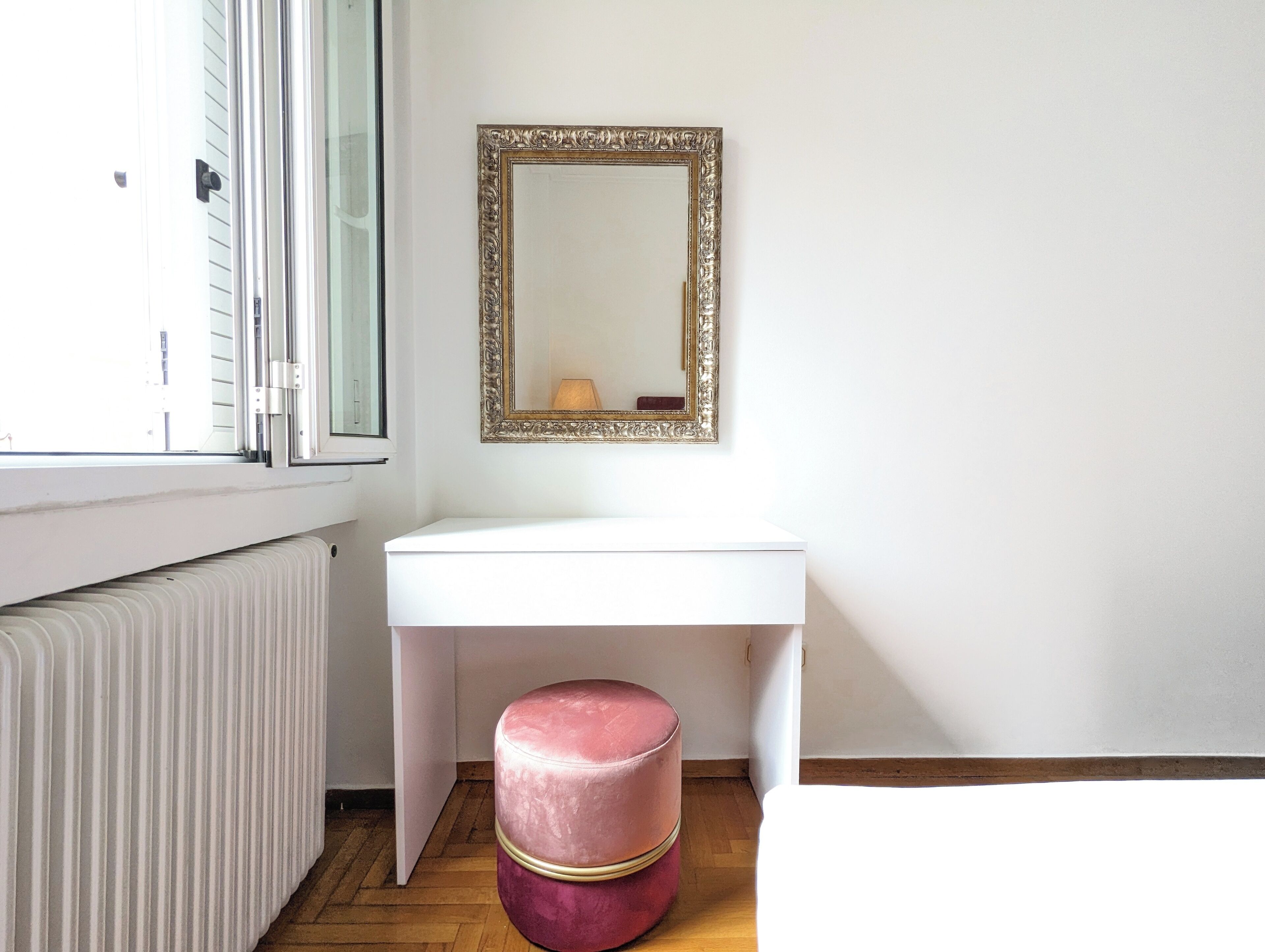Een witte badkamer met een radiator, een witte wasbak, een roze kruk en een spiegel met een gouden lijst.