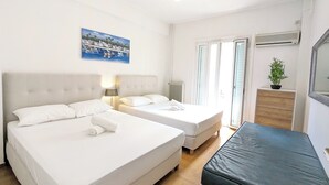 Quarto de hotel com duas camas, chão de madeira, sofá preto, janela com persianas, radiador, ar condicionado fixado na parede, quadro emoldurado e planta em vaso.