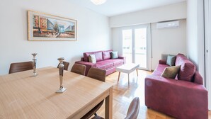 A modern living room with a wooden dining table, two sofas, and a large window with a view of buildings. - Athens Crown Paradise Apartments (Athens)