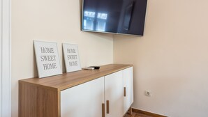 A modern living room with a flat-screen TV mounted on the wall, a wooden cabinet with two framed signs, and a white wall. - Athens Crown Paradise Apartments (Athens)