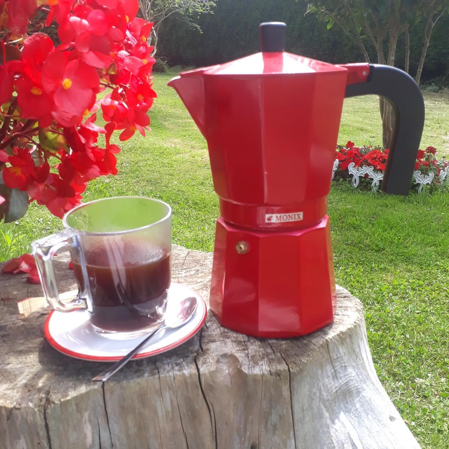 Uma cafeteira vermelha sobre um toco de madeira, com uma chávena de vidro de café e uma colher ao lado.