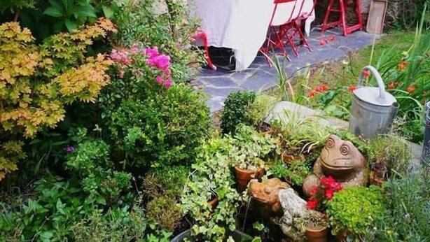 A rustic outdoor dining area with a stone wall, a table covered with a white tablecloth, red chairs, and potted plants. - A Cortiña de Canalarrana (Ribadeo)