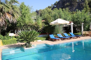 A pool area with lounge chairs and an umbrella, surrounded by greenery and a mountainous backdrop. - Hotel Dalos (Fethiye)