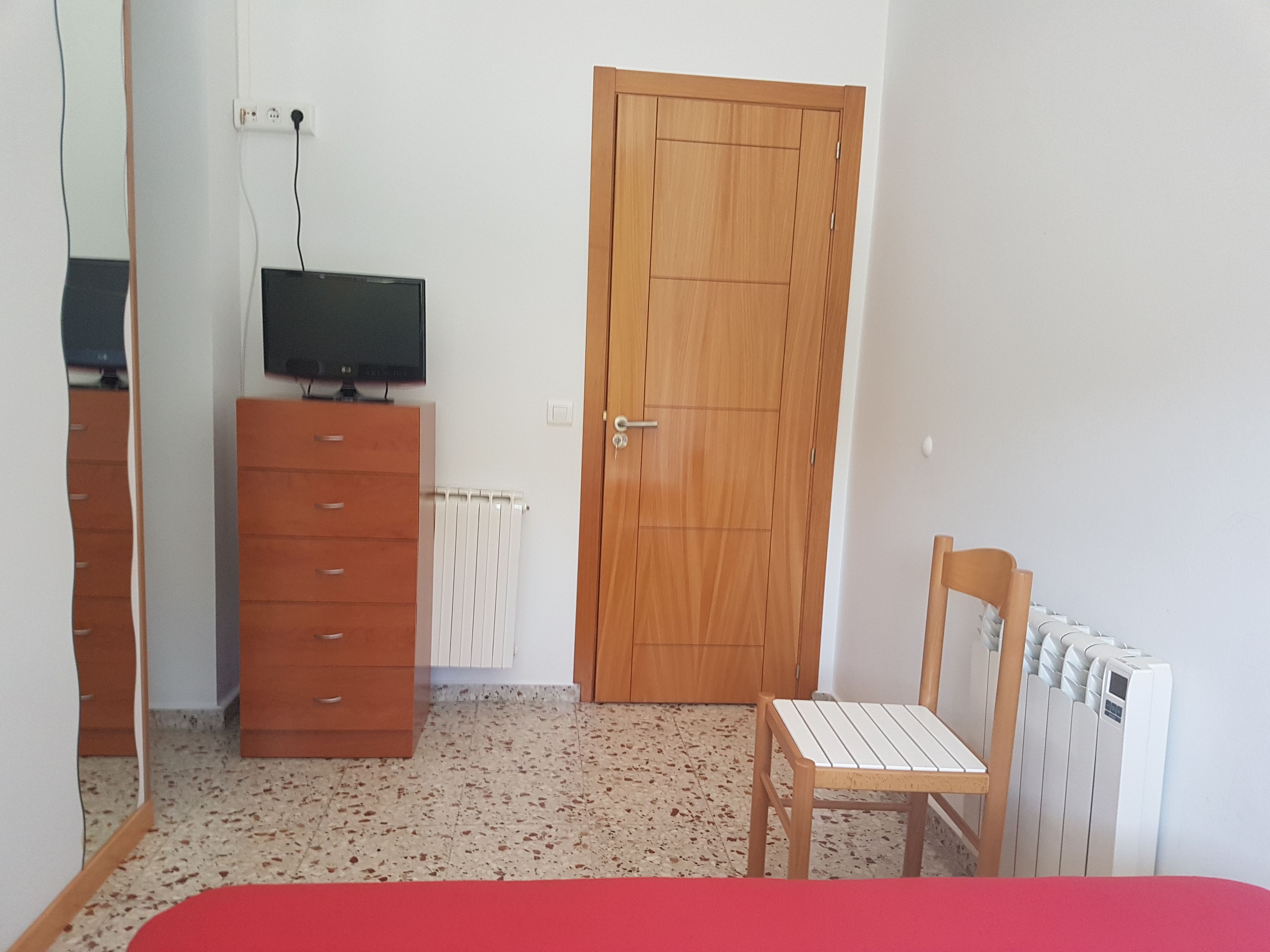 Habitació amb un armari de fusta, un televisor a sobre, una porta de fusta, un radiador i una cadira.