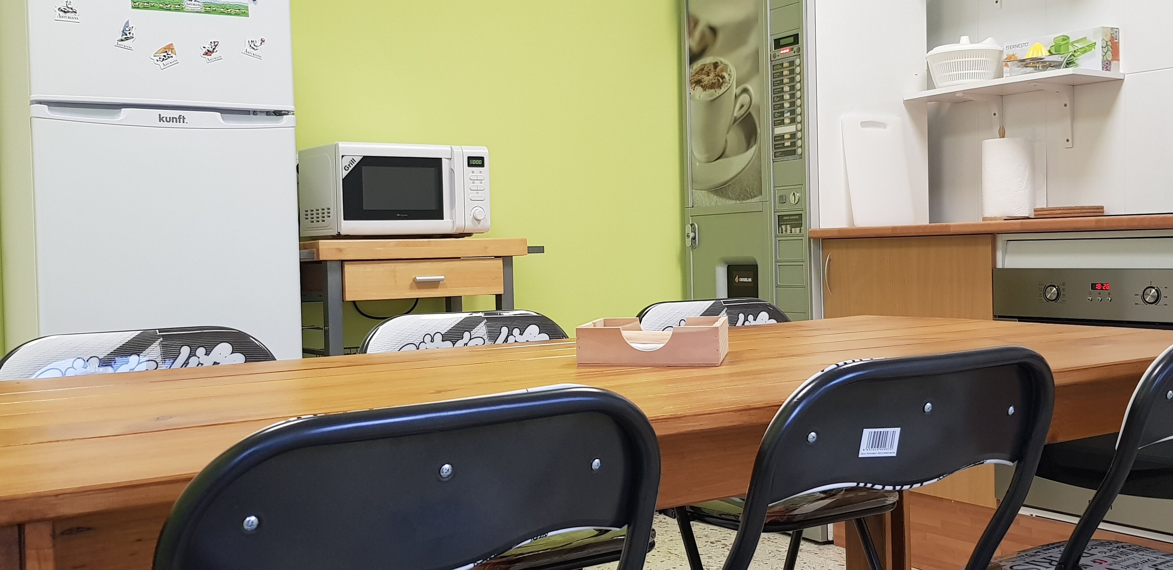 A dining area with a wooden table, black chairs, a microwave, a refrigerator, and a kitchen counter with a coffee machine.