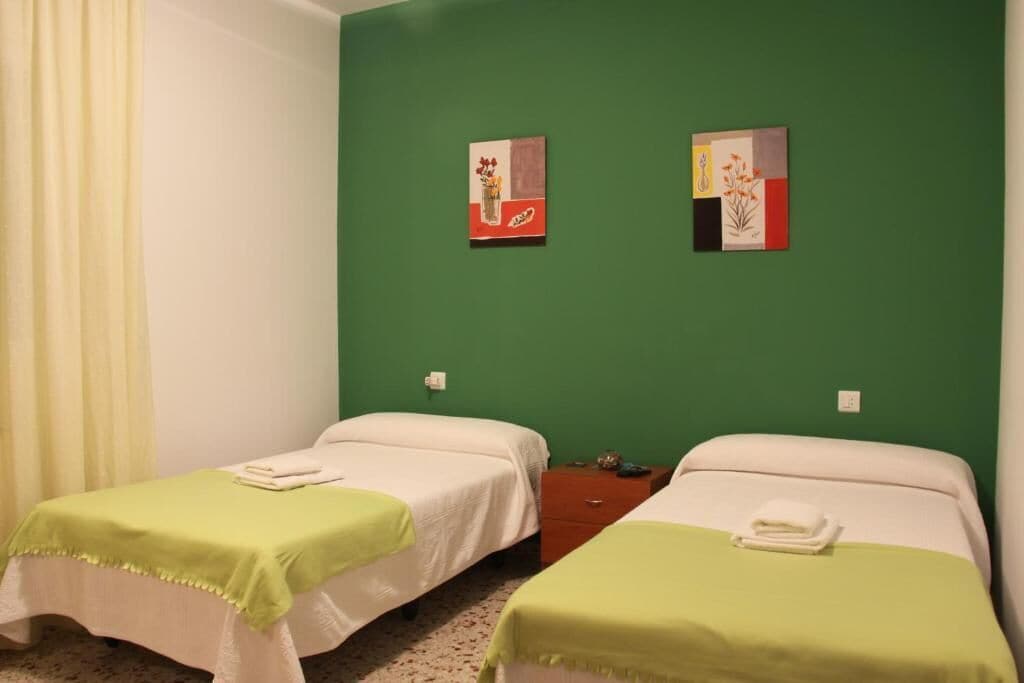 Ein Zimmer mit zwei Betten, jedes mit einer grünen Tagesdecke und weißen Bettwäsche, sowie zwei an der Wand hängenden Kunstwerken.