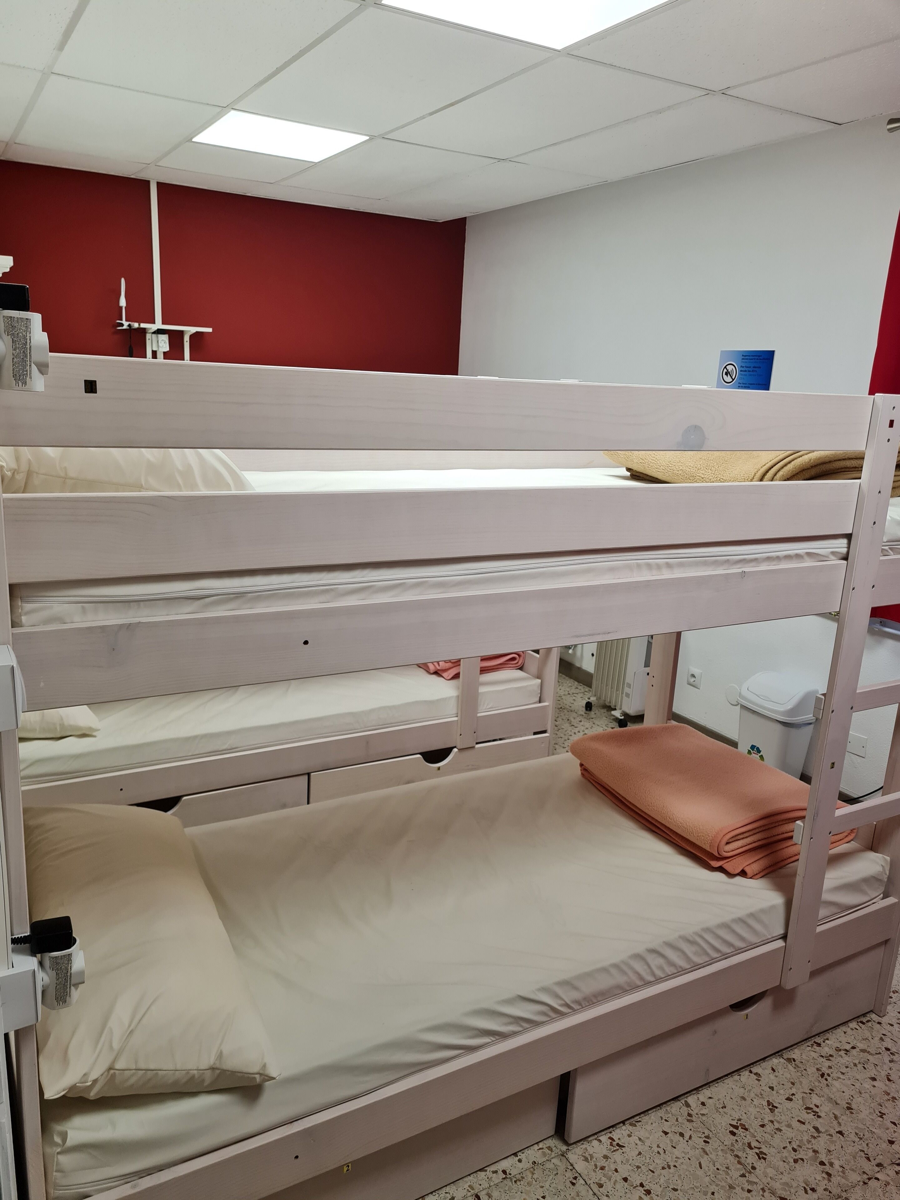 Un lit superposé à trois niveaux, un mur rouge et un plafond blanc avec des néons.