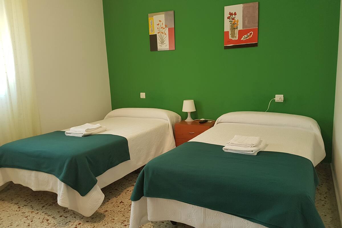 Une chambre avec deux lits, chacun recouvert d’une couverture verte et agrémenté d’oreillers blancs, une table de chevet avec une lampe, et deux œuvres d’art murales.