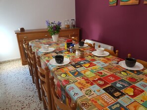 Uma mesa de jantar com toalha de mesa colorida, cadeiras de madeira e diversos objetos sobre a mesa, incluindo xícaras, um vaso com flores e frascos de condimentos.