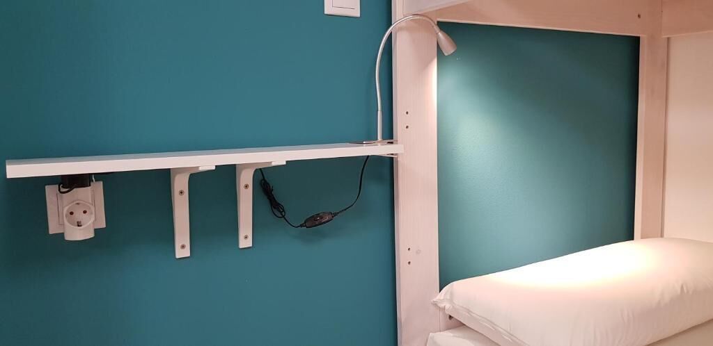 Una cama litera con colchón blanco y pared verde azulado. Hay una estantería blanca con toma de corriente y un cable.