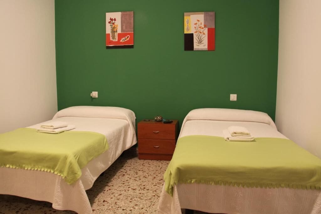 A room with two beds, each with a green blanket and white bedding. There are two paintings on the wall above the beds.