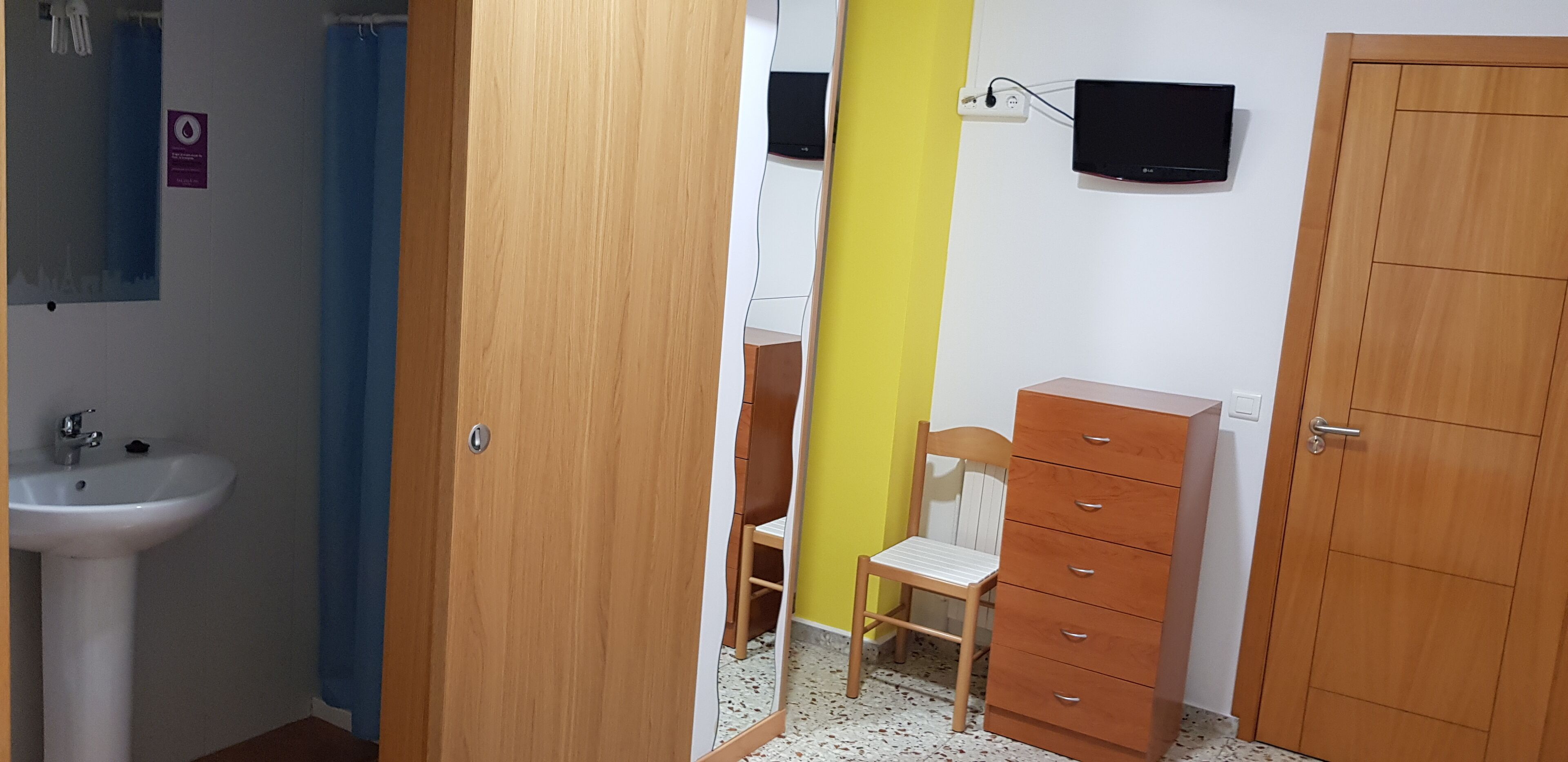 Una habitación con un armario de madera, una silla, un mueble pequeño, un televisor montado en la pared y un lavabo.