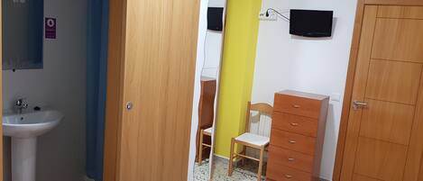 Una habitación con un armario de madera, una silla, un mueble pequeño, un televisor montado en la pared y un lavabo.