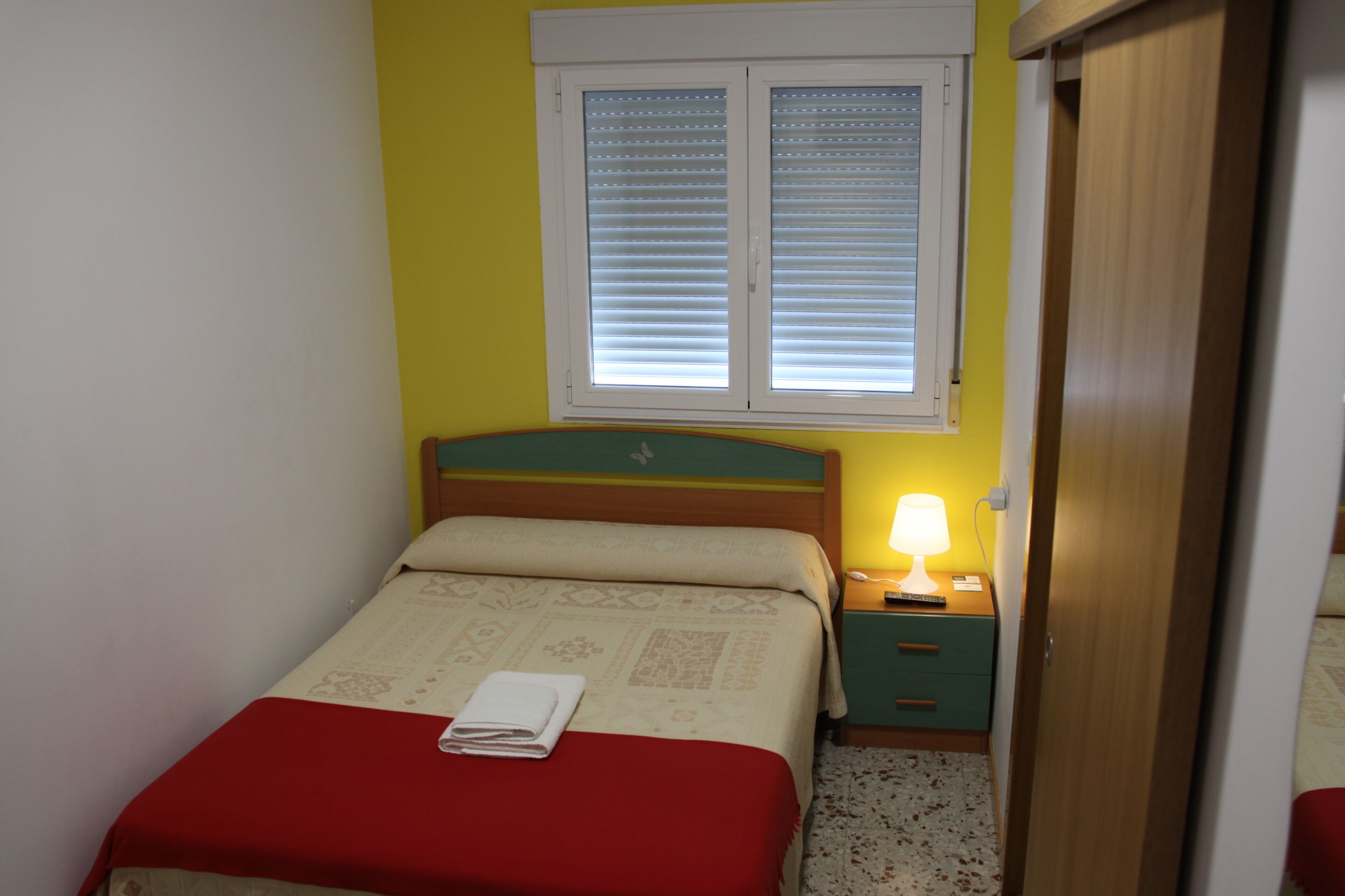 Un dormitorio con cama, mesita de noche, lámpara y ventana con persianas.