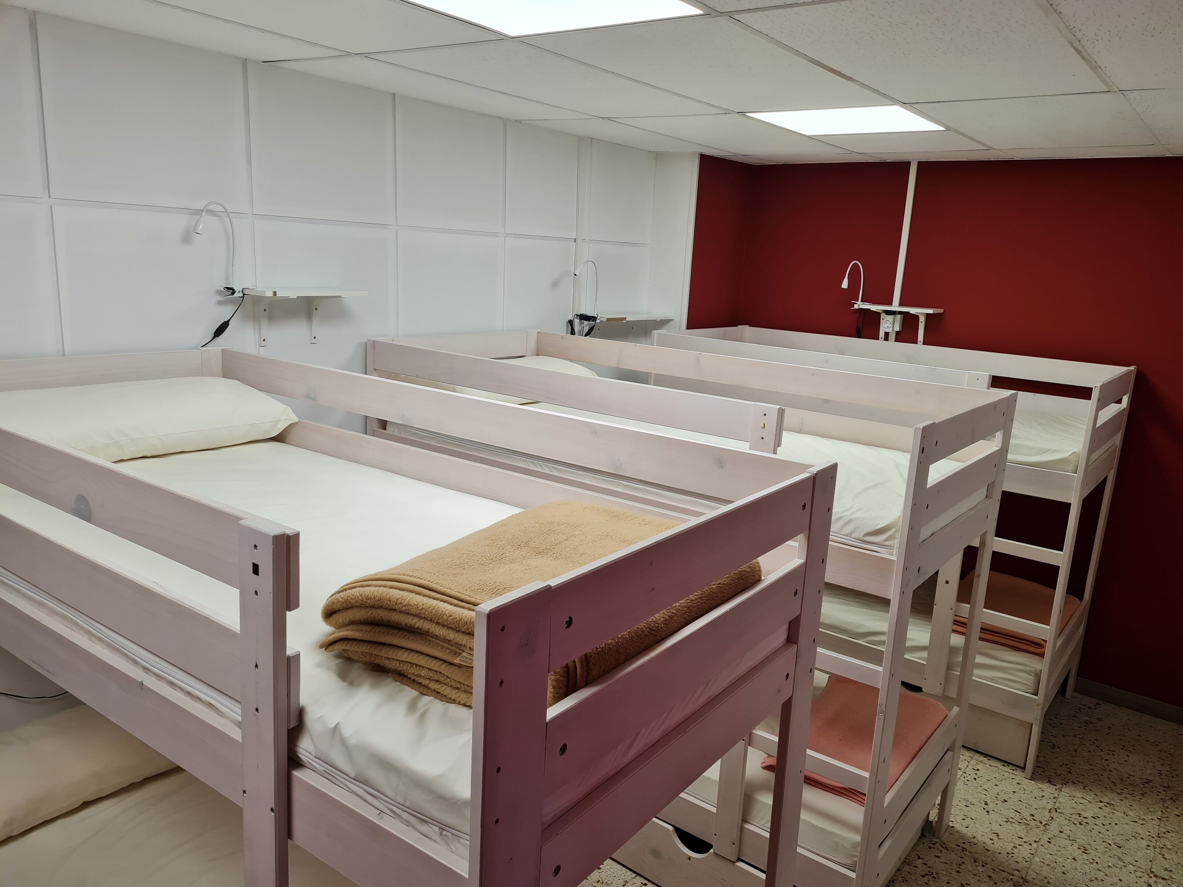 Ein Zimmer mit vier Etagenbetten, jeweils mit einer weißen Matratze und einer beige farbenen Decke. Eine Wand ist rot, die Decke weiß mit Neonbeleuchtung.