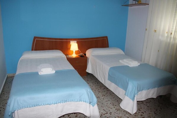 Dos camas individuales con ropa de cama azul y blanca, un cabecero de madera, una mesita de noche con lámpara y un armario blanco.