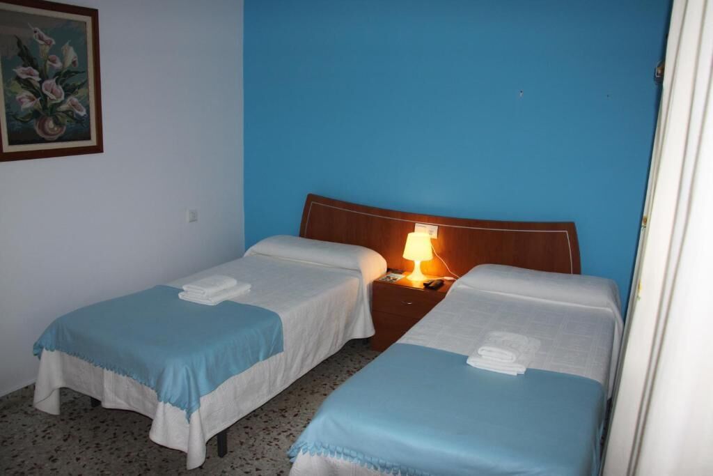 A hotel room with two single beds, a wooden headboard, a nightstand with a lamp, and a painting on the wall.