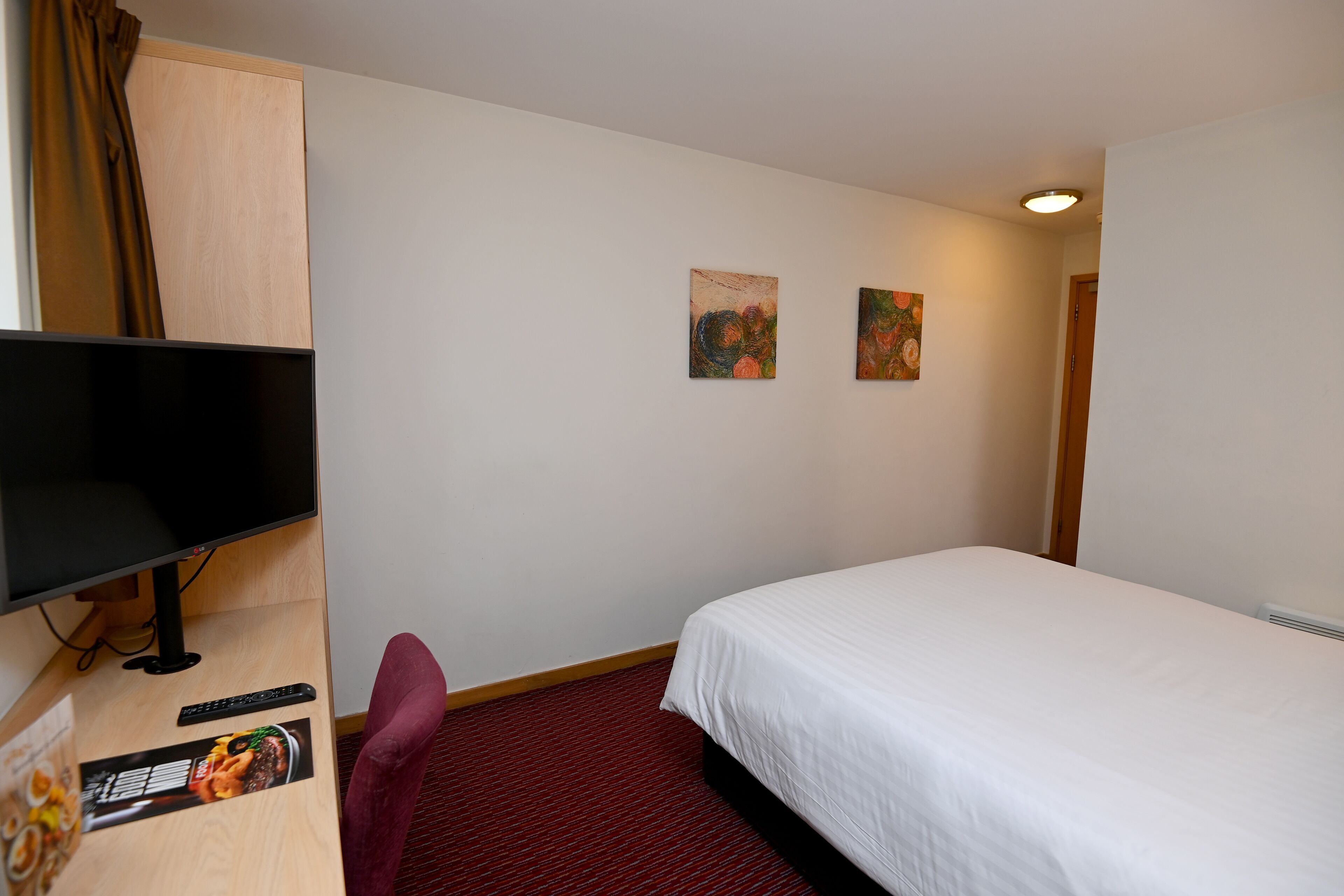 Een hotelkamer met een bed, een bureau met een computerscherm, een stoel en twee schilderijen aan de muur.