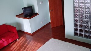 A room with a wooden floor, a red sofa, a television on a shelf, a door, and a window with a grid pattern. - Motel Abalo (Catoira)