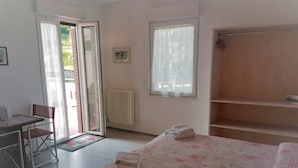 Un dormitorio con una cama, un escritorio y una silla. Se observa un radiador, una ventana y una puerta que da a un espacio al aire libre.