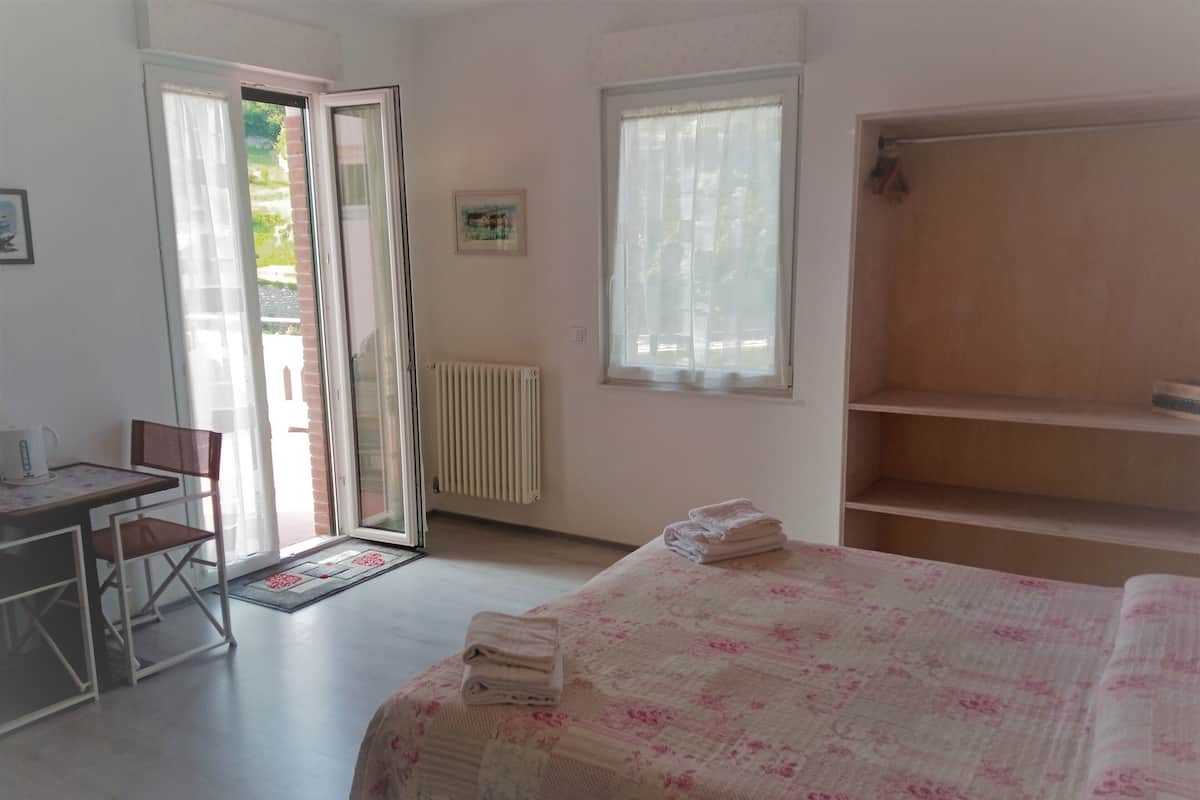 A bedroom with a bed, a desk, and a chair. There is a radiator, a window, and a door leading to an outdoor area.
